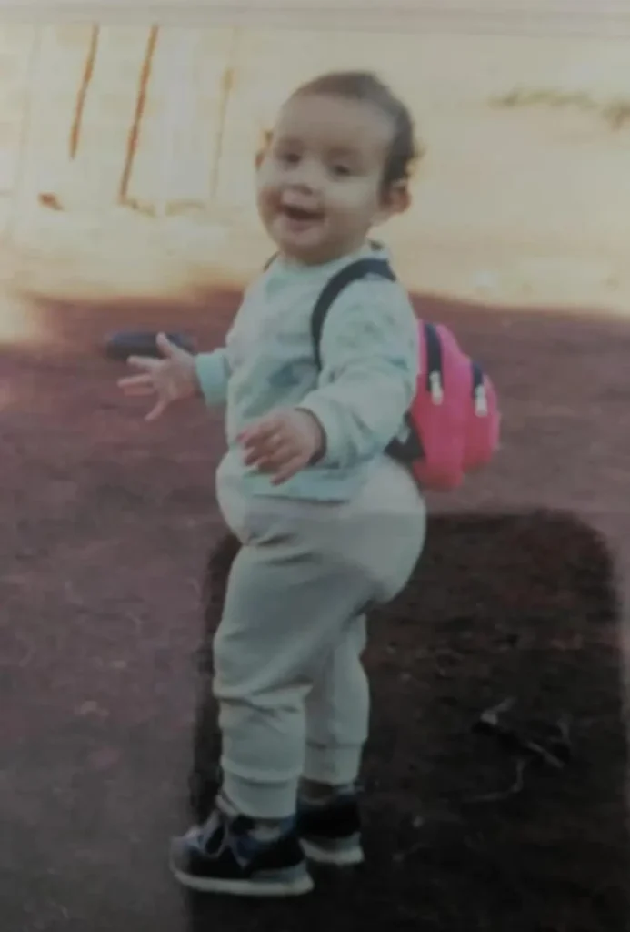 Antiga foto de infância da Psicóloga Jéssica Mattos quando bebê, em pé com os braços abertos equilibrando e uma expressão curiosa e surpresa. O bebê veste uma camisa verde-clara, calças beges e uma pequena mochila rosa. Imagem utilizada para humanizar a página Sobre do consultório online.